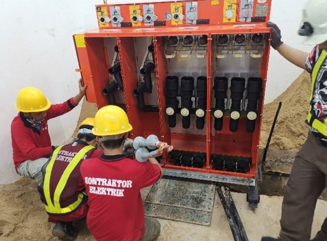 Workers in yellow hard hats and red shirts kneeling at an open orange feeder pillar with cable terminations