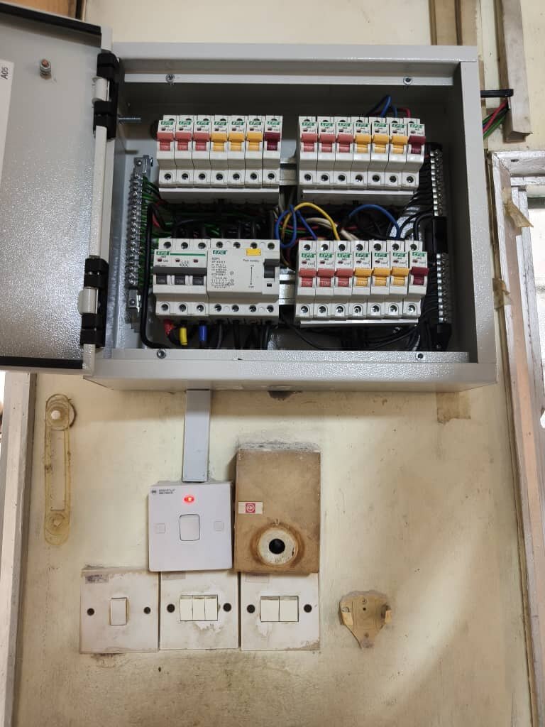 Open grey metal distribution board with a main switch, a residual current circuit breaker and three groups of miniature circuit breakers marked red, yellow and blue, above old switches and a doorbell transformer
