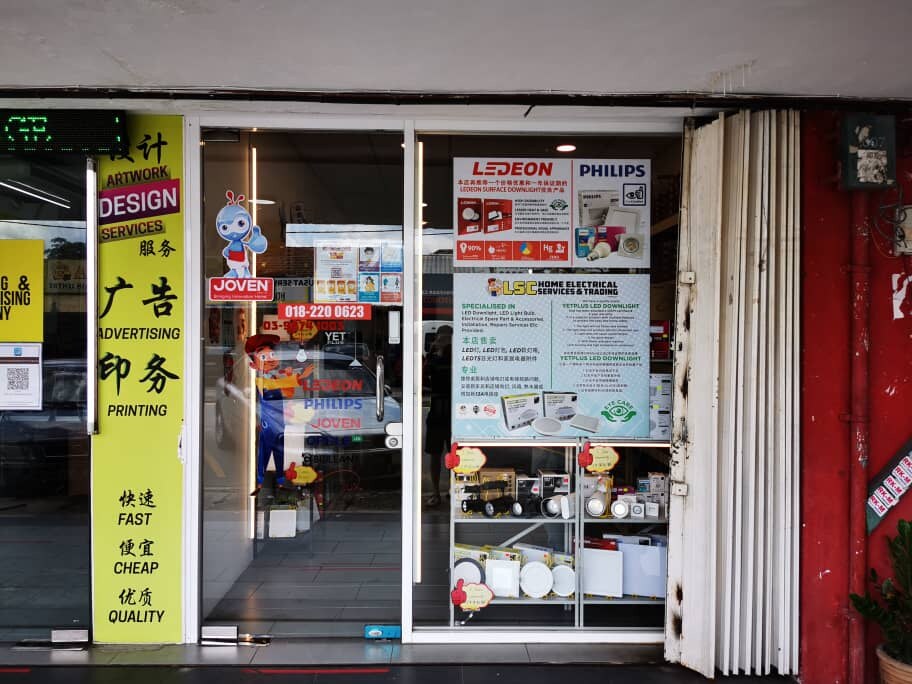 Glass shop door with a large poster for Ledeon and Philips LED lights, an LSC Home Electrical poster and phone-number stickers, with boxed downlights and track lights on a display shelf behind