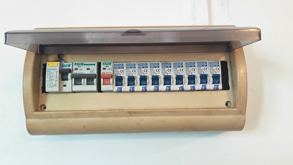 Beige surface-mounted distribution board with its clear cover raised, showing an RCCB, a double-pole main switch, a red 10A breaker and nine blue Berlin miniature circuit breakers