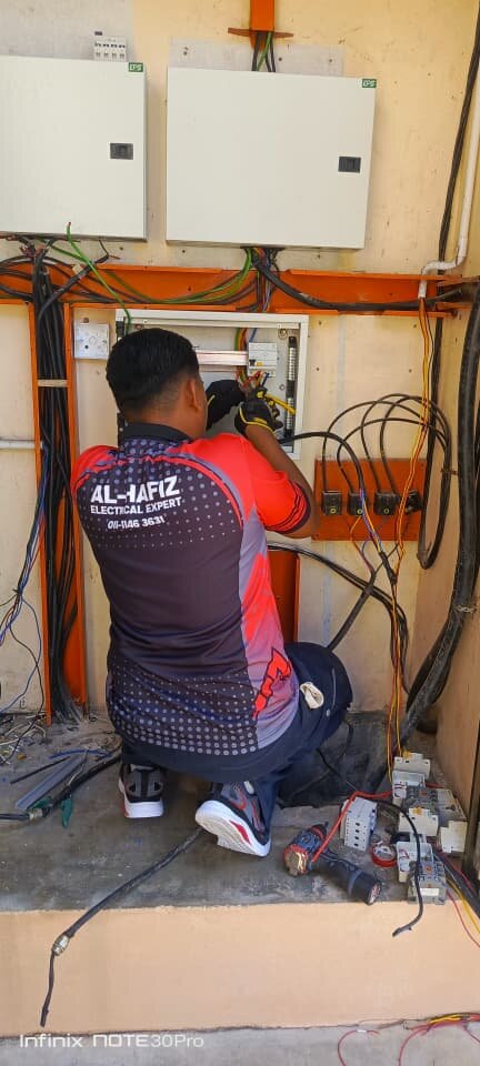 Electrician kneeling at a small open distribution board under two larger grey enclosures, with cables on orange trunking; his shirt reads Al-Hafiz Electrical Expert 011-1146 3631