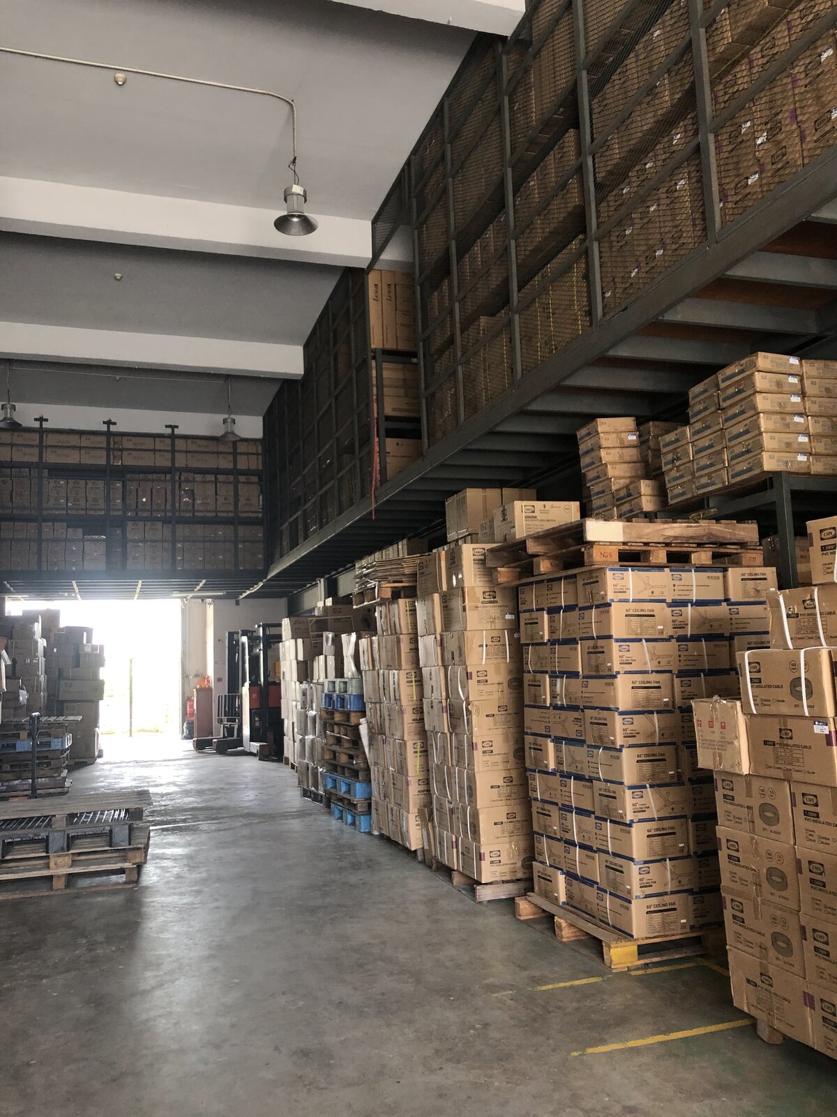 Warehouse aisle with tall stacks of cardboard cartons on pallets, steel racking above holding more cartons, and a bright loading door at the far end