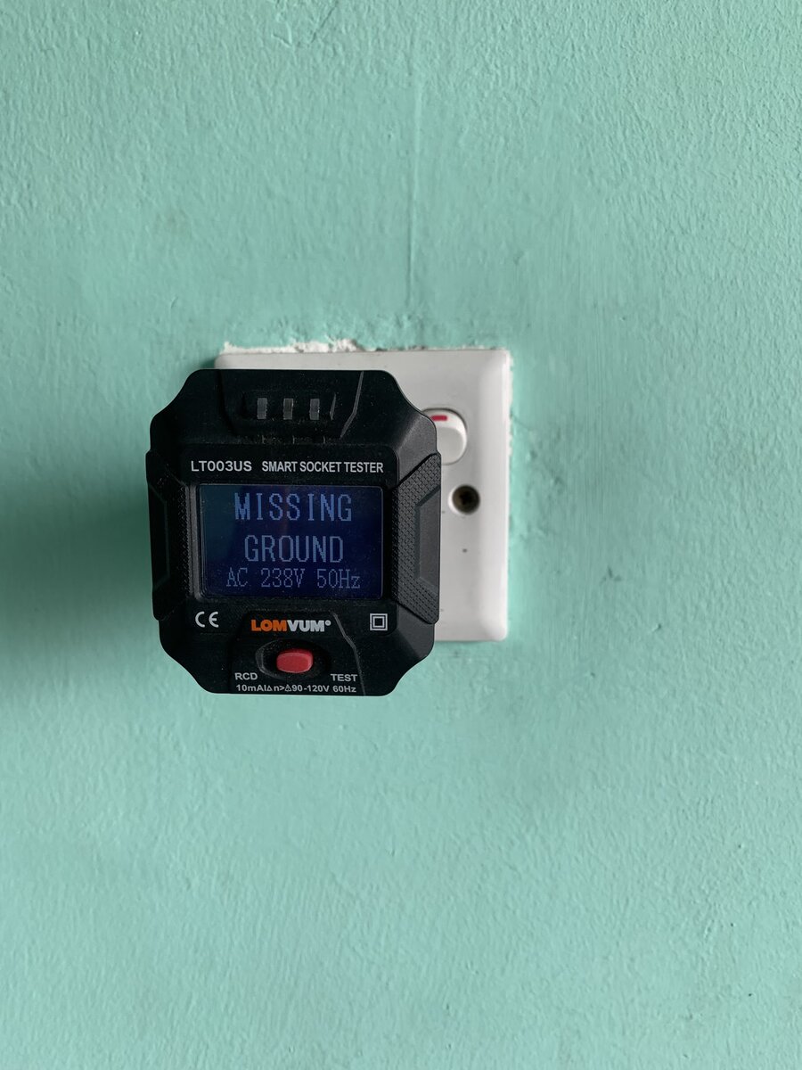 Black plug-in socket tester in a white wall socket on a mint-green wall, its screen reading MISSING GROUND, AC 238V 50Hz