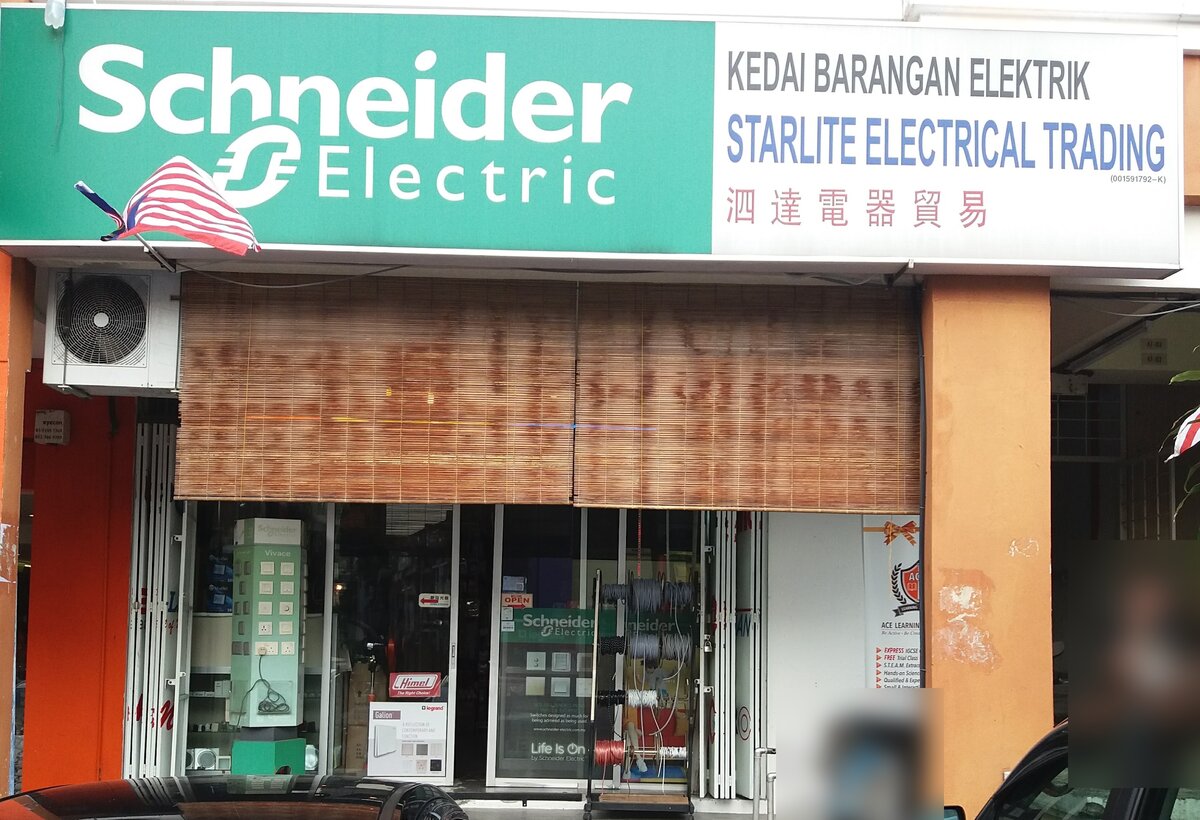 Shopfront with a green Schneider Electric sign and a white Starlite Electrical Trading signboard above bamboo blinds; behind the glass a green switch display tower, a red Himel sticker, a Legrand poster and a Schneider Life Is On poster, and a rack of wire reels by the door