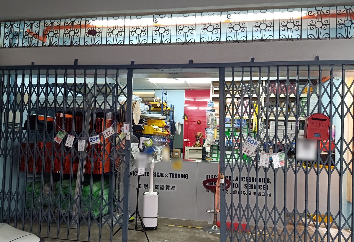 Shop interior seen through a folding metal grille: a grey counter lettered E Nice Electrical & Trading and Electrical Accessories, Installation Services, orange trunking on racks to the left, cable drums, yellow parts bins and shelves of boxes