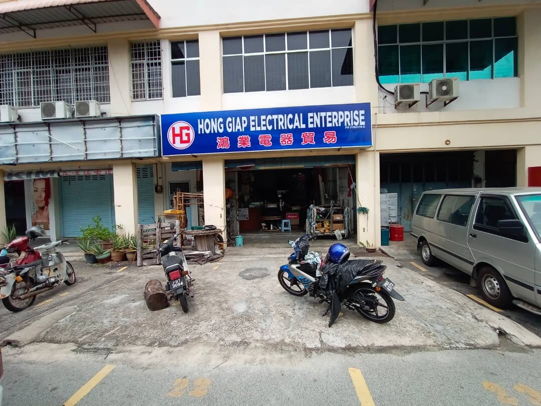 Two-storey shop row with a blue 'HG Hong Giap Electrical Enterprise 鴻業電器貿易' signboard over an open shopfront, motorcycles and a van parked in front