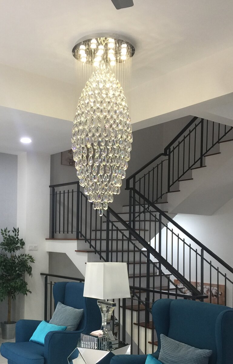 Tall teardrop-shaped crystal chandelier hanging in a double-height stairwell above blue armchairs and a table lamp