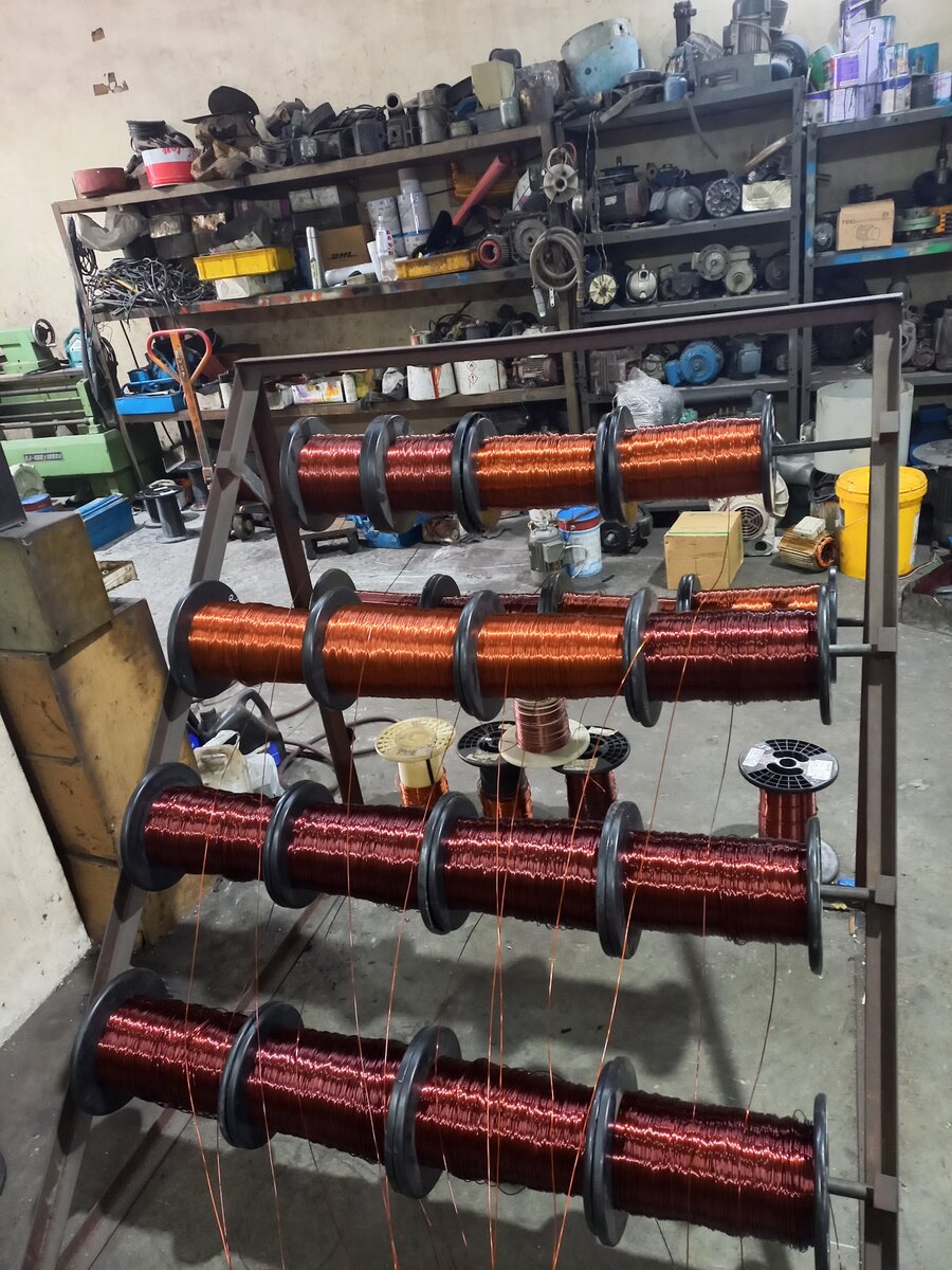 Metal rack holding four rows of copper winding-wire reels in a workshop with shelves of motor parts behind