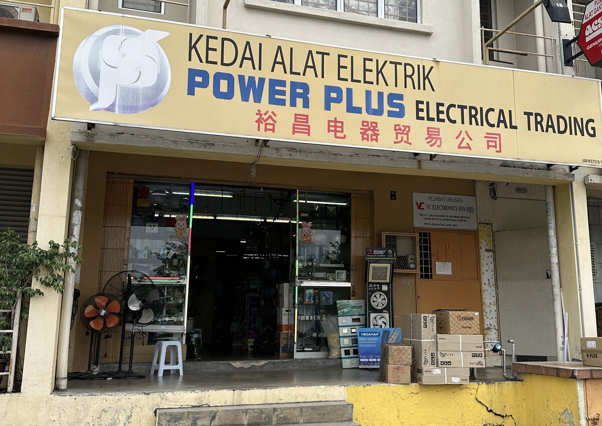 Shop front with a pale yellow Power Plus Electrical Trading signboard, stand fans, a switch display stand and stacked cartons