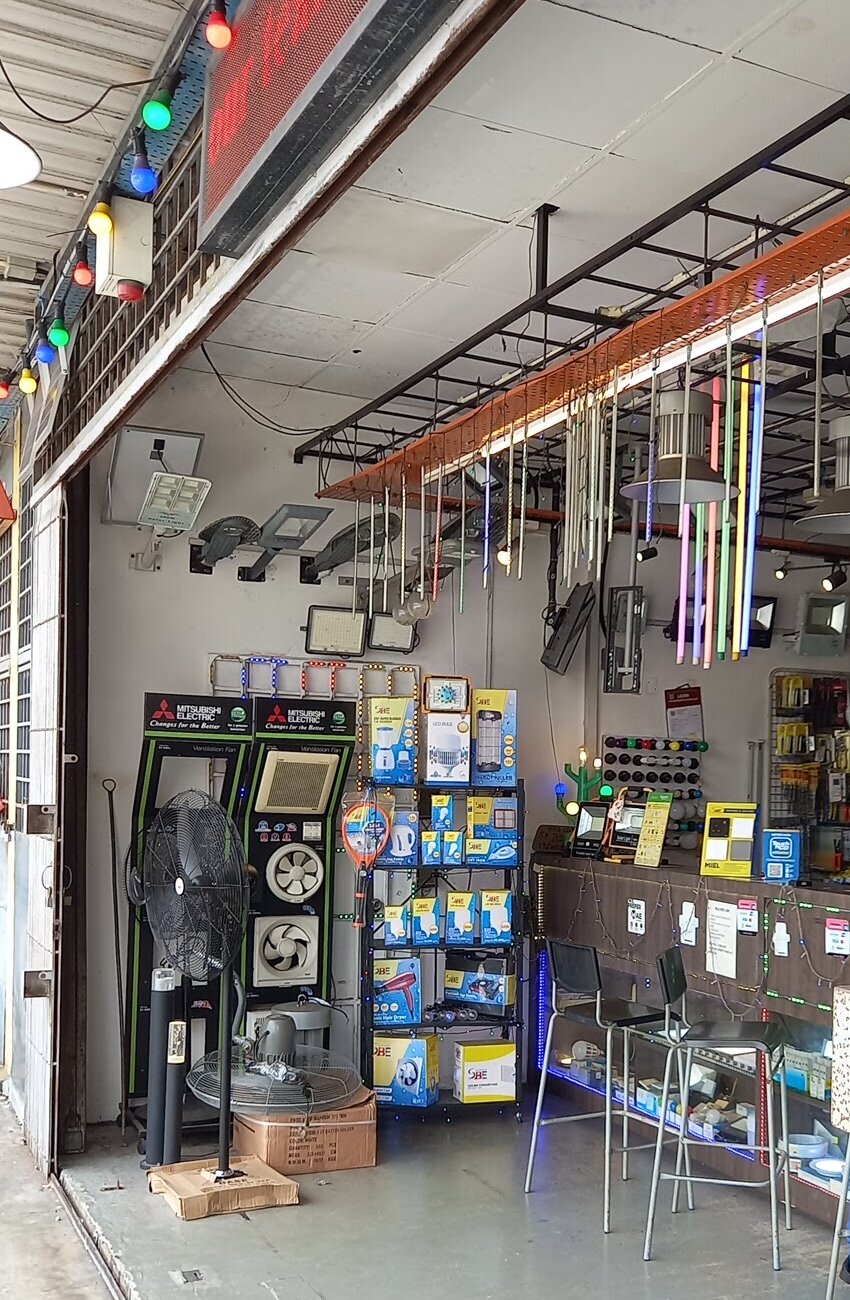 Shop entrance with two black ventilation-fan display stands, a stand fan, a shelf of blue and yellow product boxes and LED tubes hanging from the ceiling