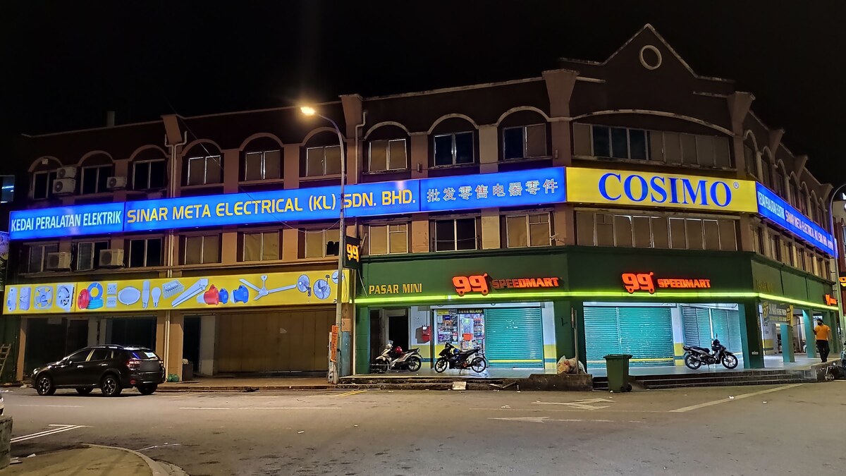 Three-storey corner shophouse block at night with a long lit blue signboard reading Sinar Meta Electrical (KL) Sdn. Bhd., a yellow COSIMO panel on the corner, a yellow strip of fan, cable and lamp pictures over closed shutters, and a 99 Speedmart on the ground floor