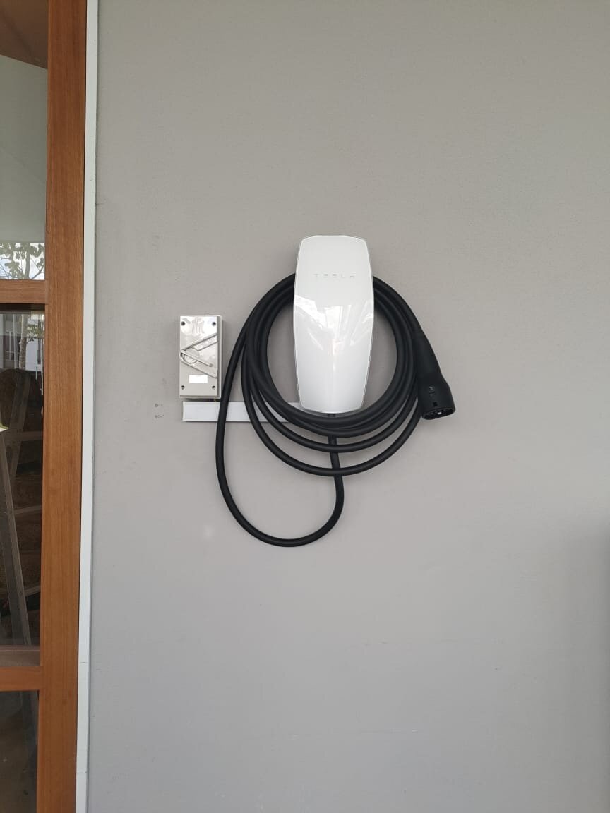 White wall-mounted electric vehicle charger with its cable coiled on the holder, beside a small wall box