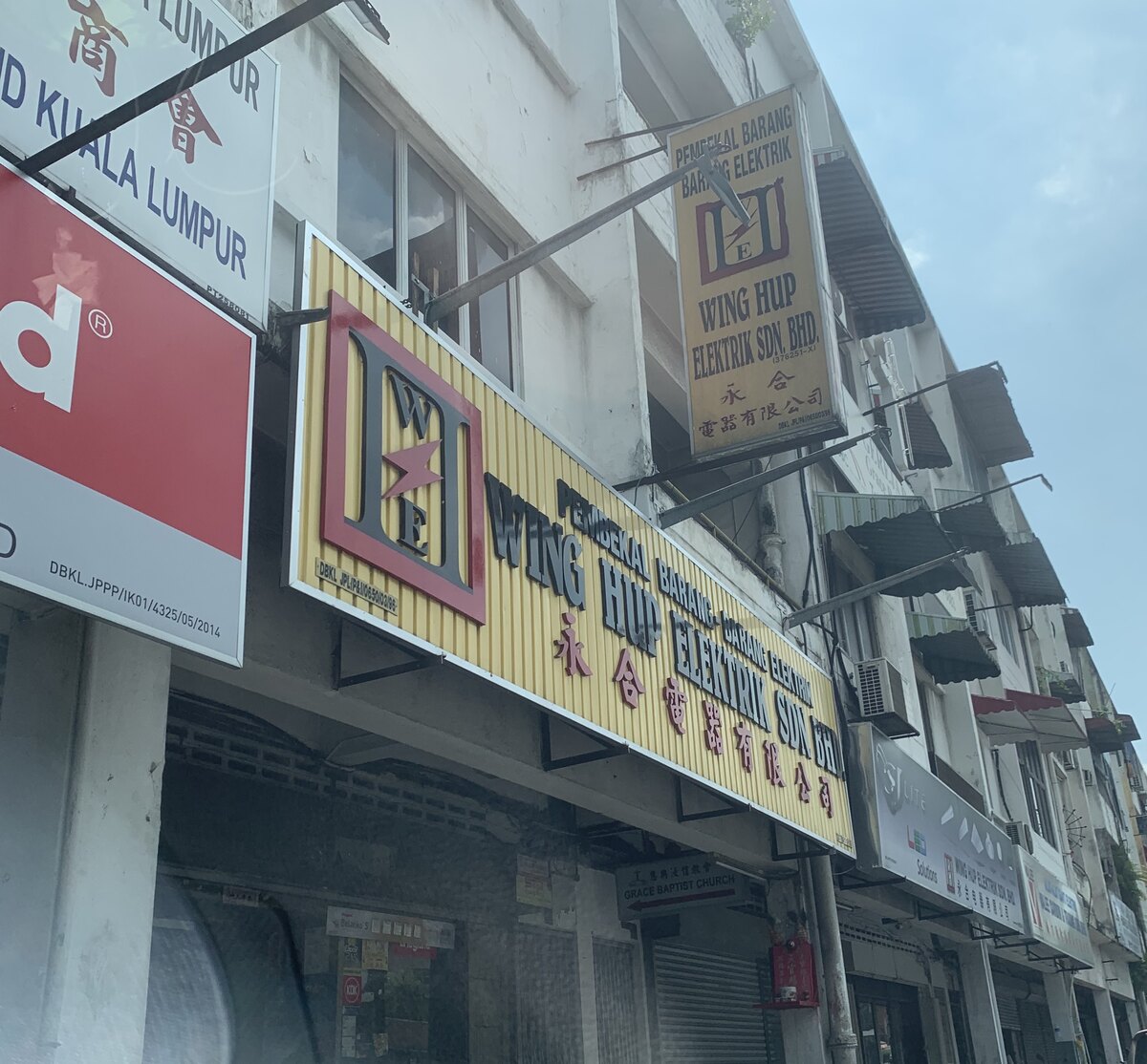 Yellow fascia sign with a logo of dark W and E letters in a red frame with a red lightning bolt and the words Wing Hup Elektrik Sdn Bhd with Chinese characters, a hanging yellow sign above, on a white shophouse row