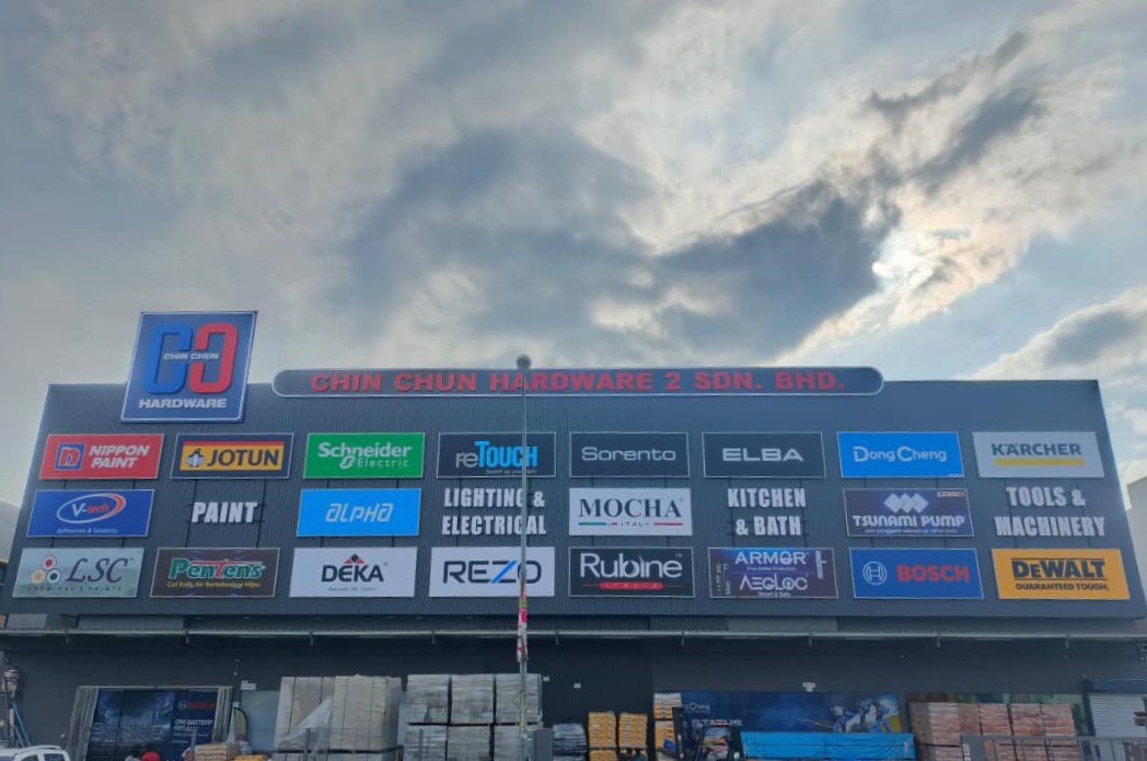 Long building facade signed Chin Chun Hardware 2 Sdn Bhd with rows of brand panels including Nippon Paint, Jotun, Schneider Electric, Bosch and DeWalt above stacked building materials