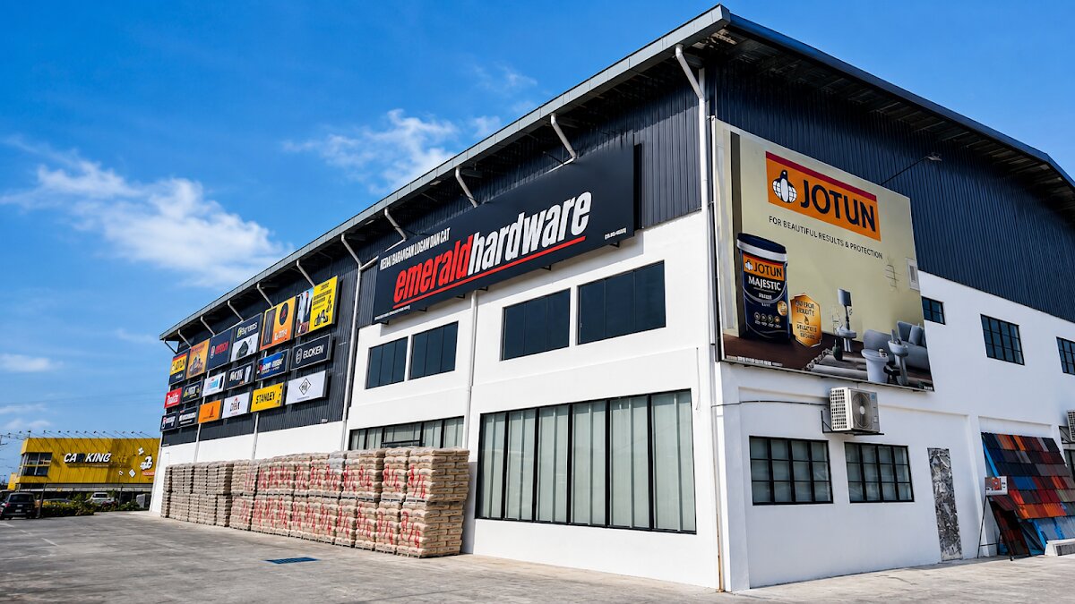Large warehouse building with a black Emerald Hardware sign, a Jotun paint billboard and stacked building materials in front