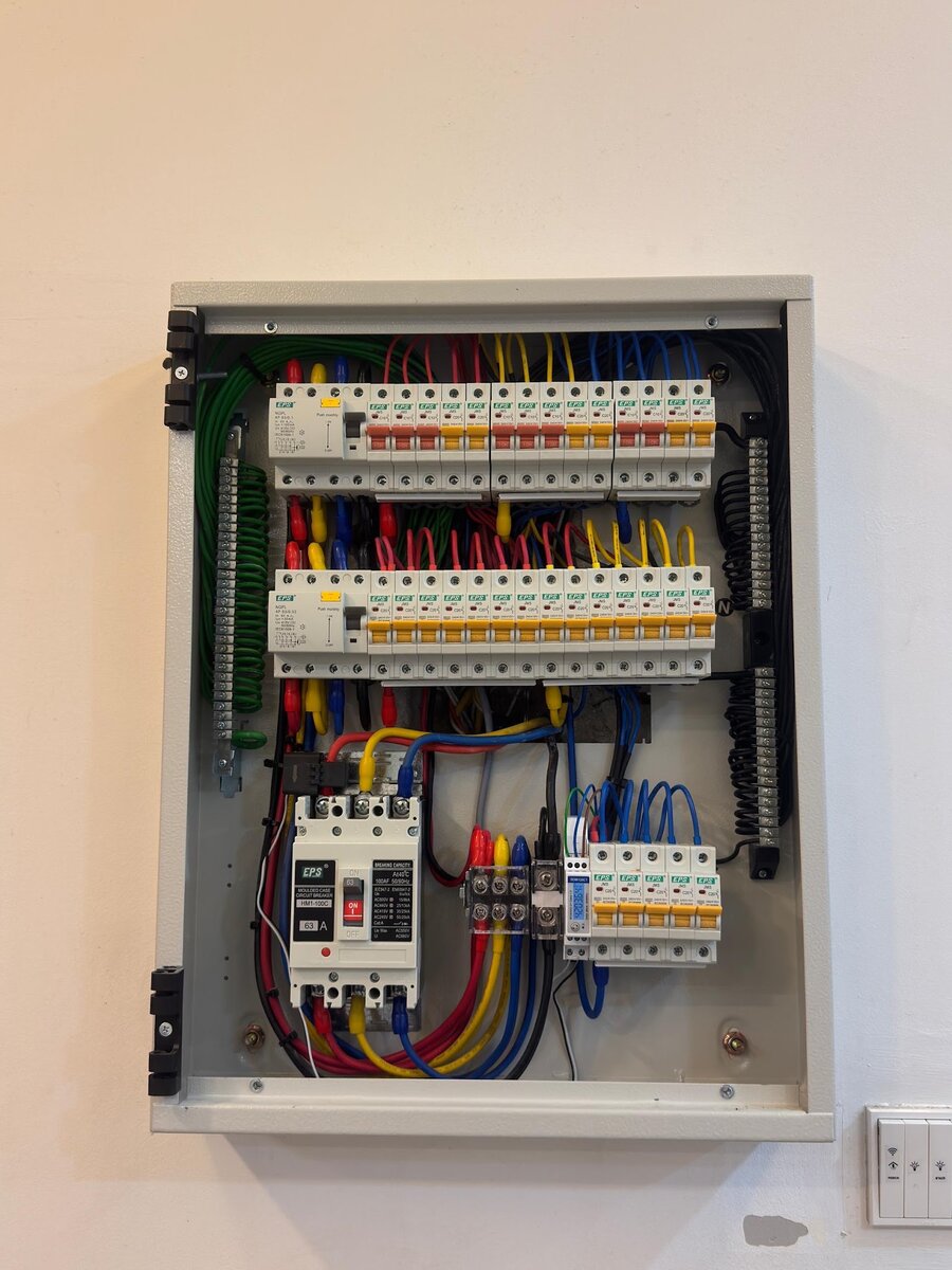 Open grey metal distribution board on a white wall with two rows of red and yellow miniature circuit breakers, a black-and-white moulded-case breaker and neatly dressed red, yellow and blue cables