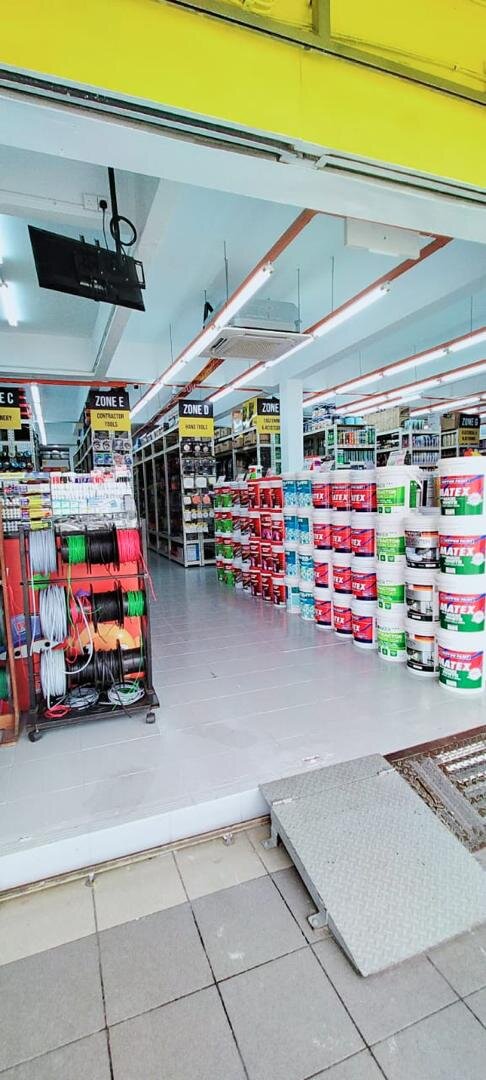 Shop entrance with a ramp, a rolling rack of wire reels on the left and stacks of paint buckets beside aisles marked by zone signs