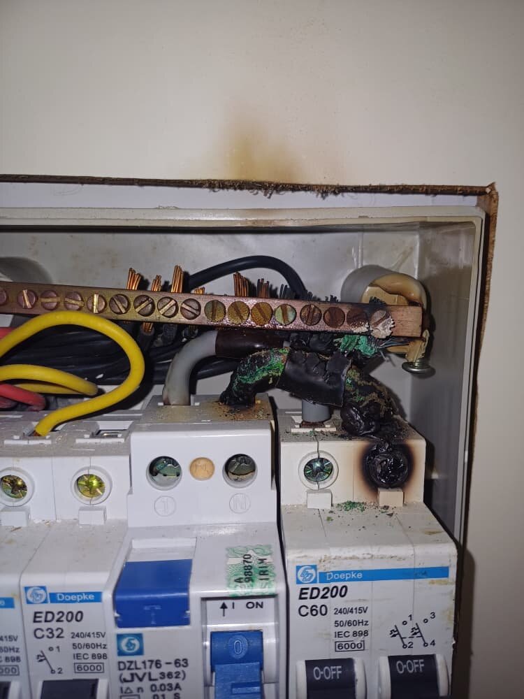 Inside a white plastic distribution board: a copper neutral bar above a row of breakers, with blackened, melted cable insulation and a burn mark on the top of the right-hand two-pole breaker and soot on the wall above