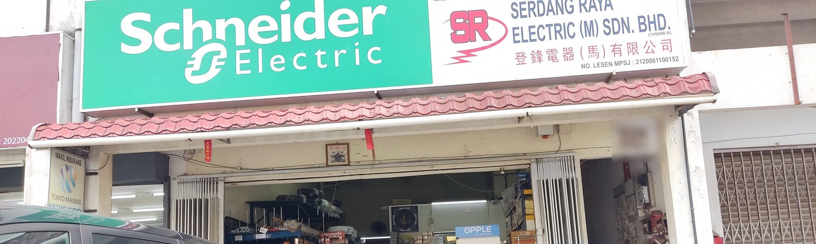 Shop front with a large green Schneider Electric sign next to a white signboard reading Serdang Raya Electric (M) Sdn. Bhd. with Chinese characters, above an open shop with shelves of electrical goods