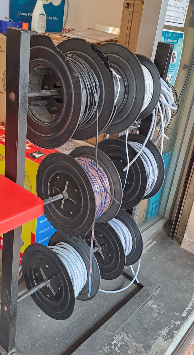 Black metal rack holding nine cable reels with grey and white flexible cable and one reel of blue and orange twin wire, by a shop doorway