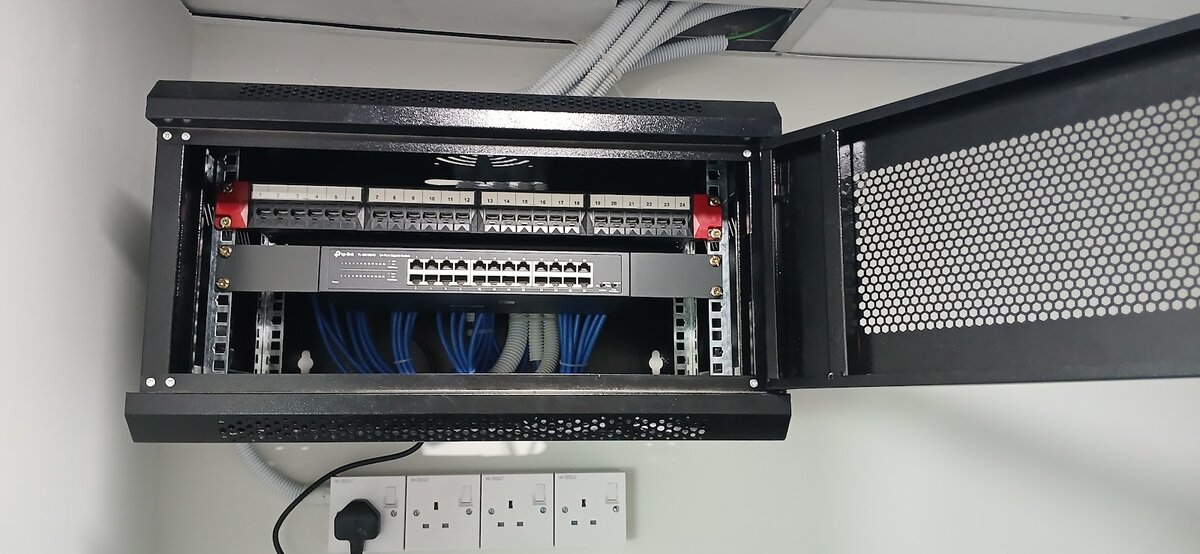 Open black wall-mounted network cabinet with a patch panel and a network switch, blue patch cables below, and four white power sockets under the cabinet