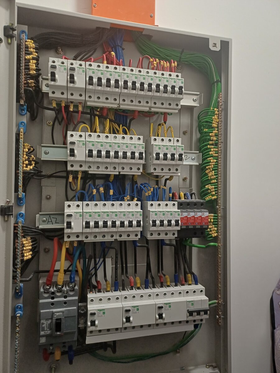 Open grey distribution board with three rows of white Schneider MCBs, red, yellow and blue cables with yellow numbered markers, a green earth bundle on the right, a red four-module surge protector at the right end of the third row, and a grey moulded-case breaker with four-pole RCCBs along the bottom