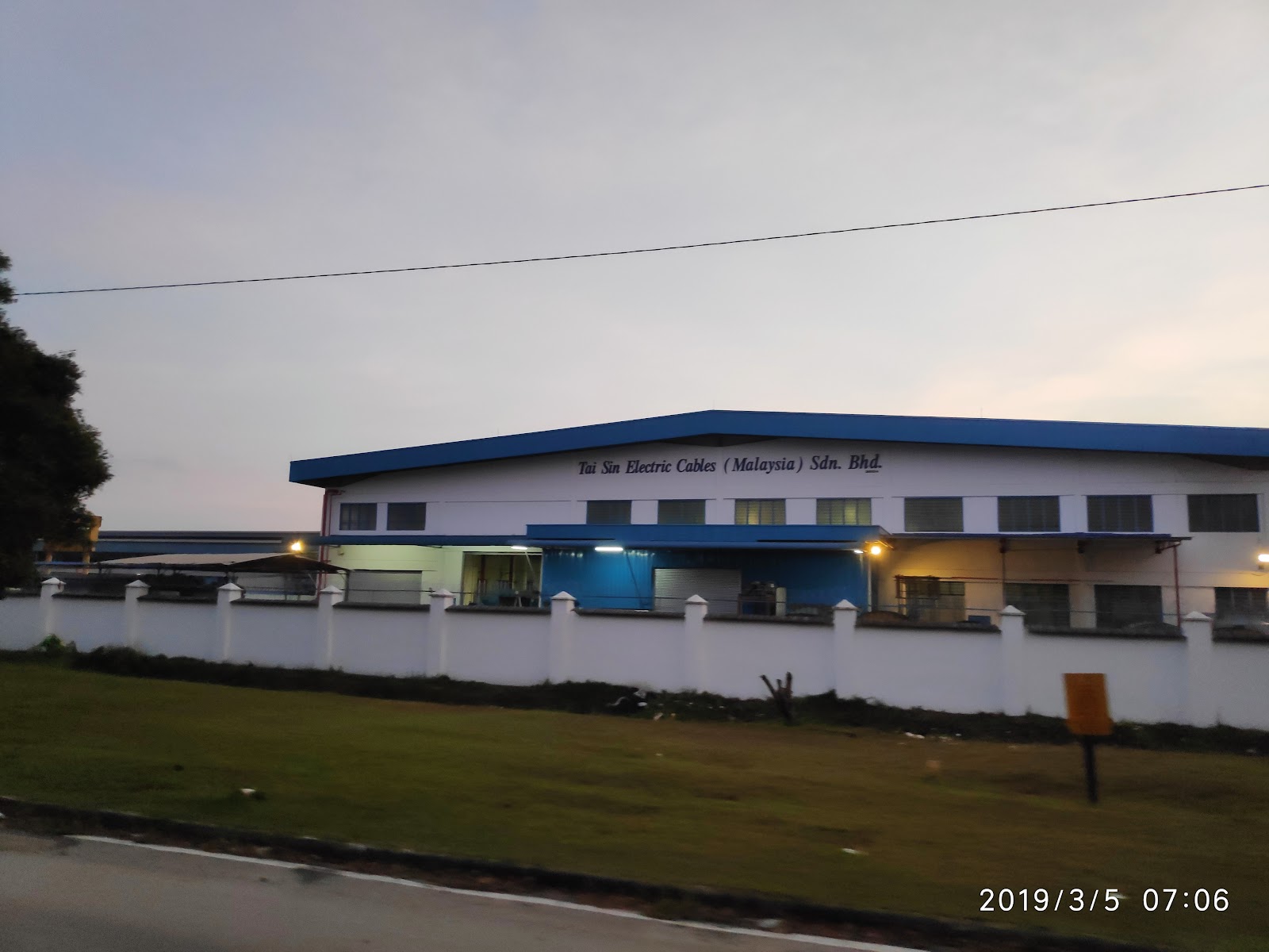 Long white factory building with a blue roof edge behind a white boundary wall