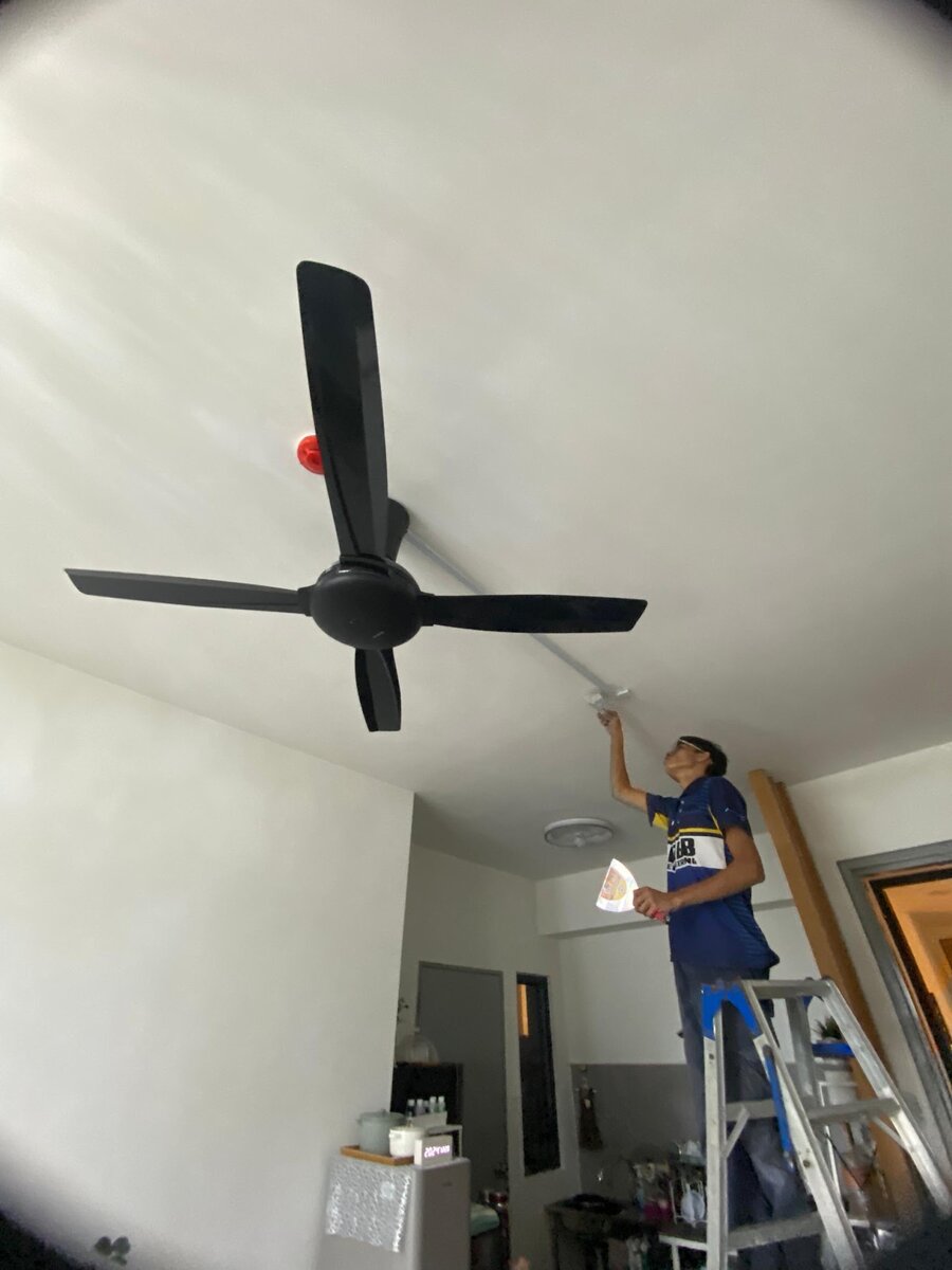 Wide-angle view of a black four-blade ceiling fan on a white ceiling, with a worker on a ladder reaching up to the ceiling beside it