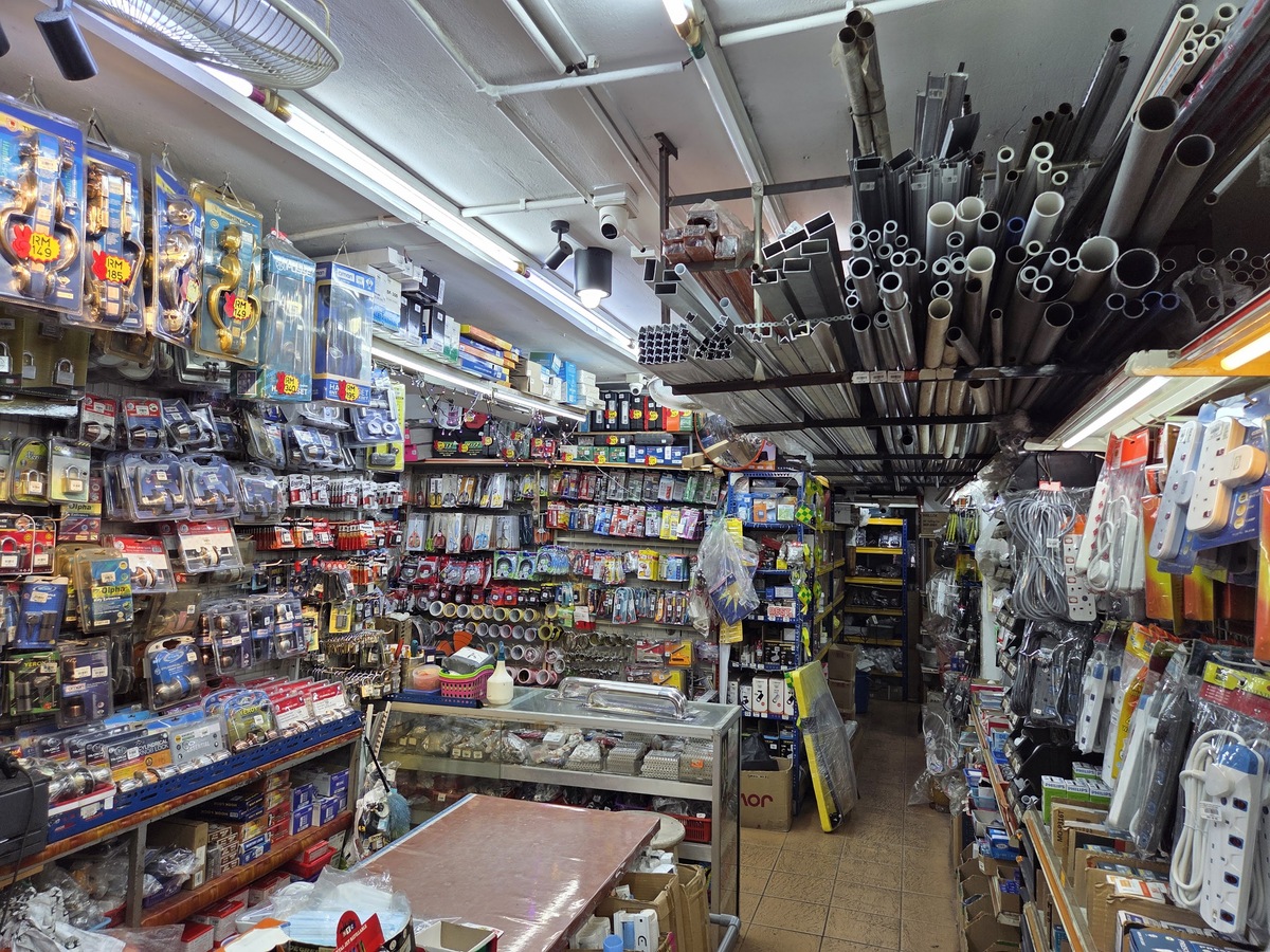 Narrow hardware shop interior with walls of packaged locks and tools, a glass counter, bundles of metal tubes on a ceiling rack and extension sockets hanging on the right