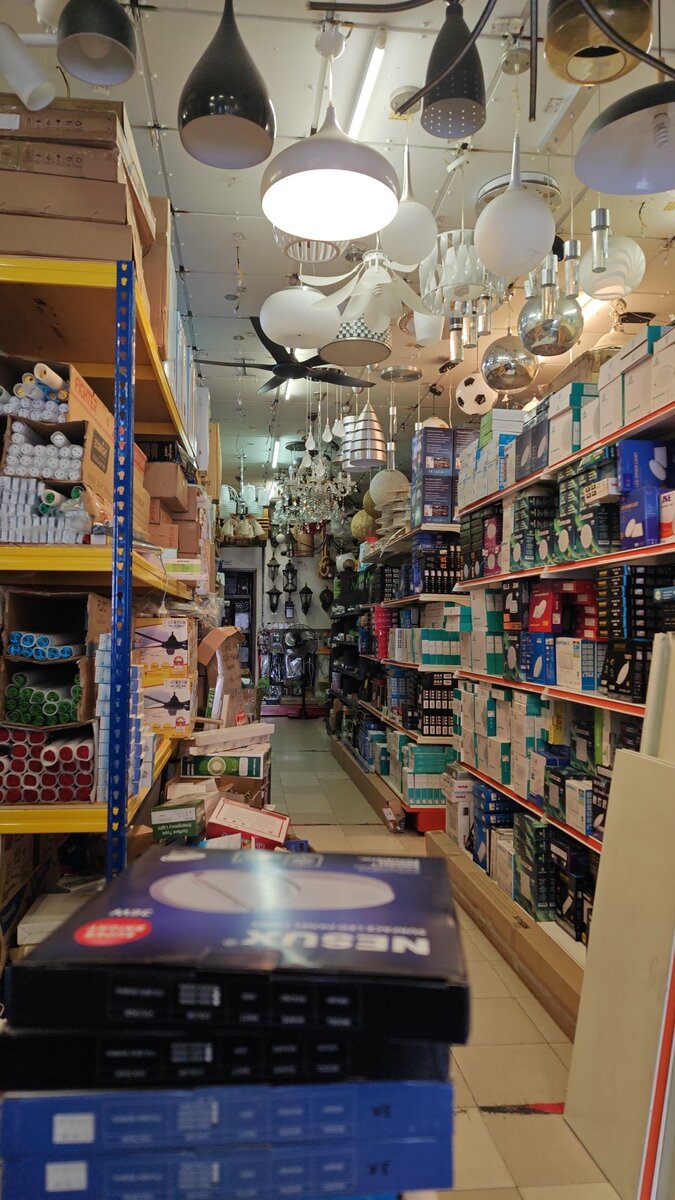 Narrow shop aisle with shelves of boxed lighting products on both sides and many pendant lamps and a ceiling fan hanging overhead