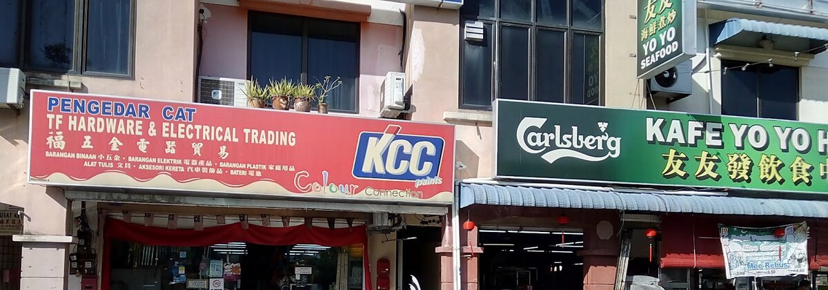 Red shop signboard reading Pengedar Cat, TF Hardware & Electrical Trading with Chinese characters and a KCC Paints logo, next to a café sign