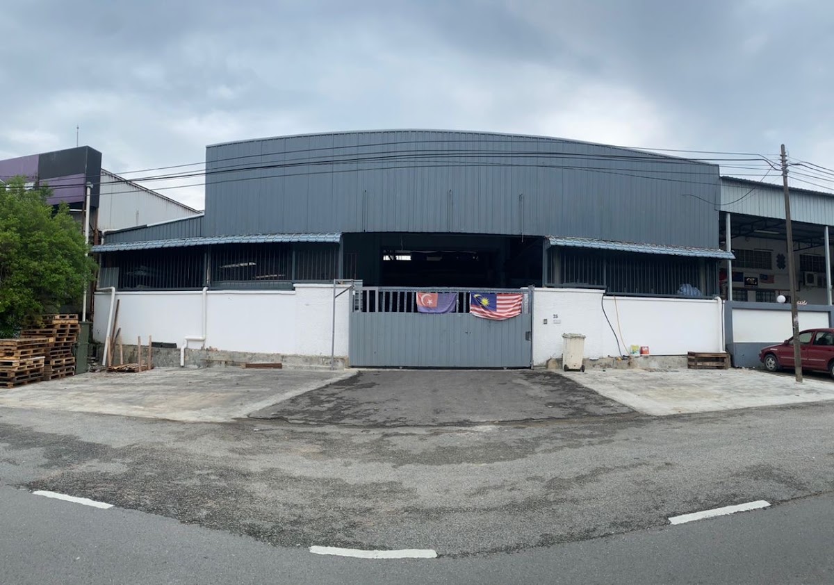 Single-storey grey warehouse behind a white wall and closed gate with Malaysian and Johor flags