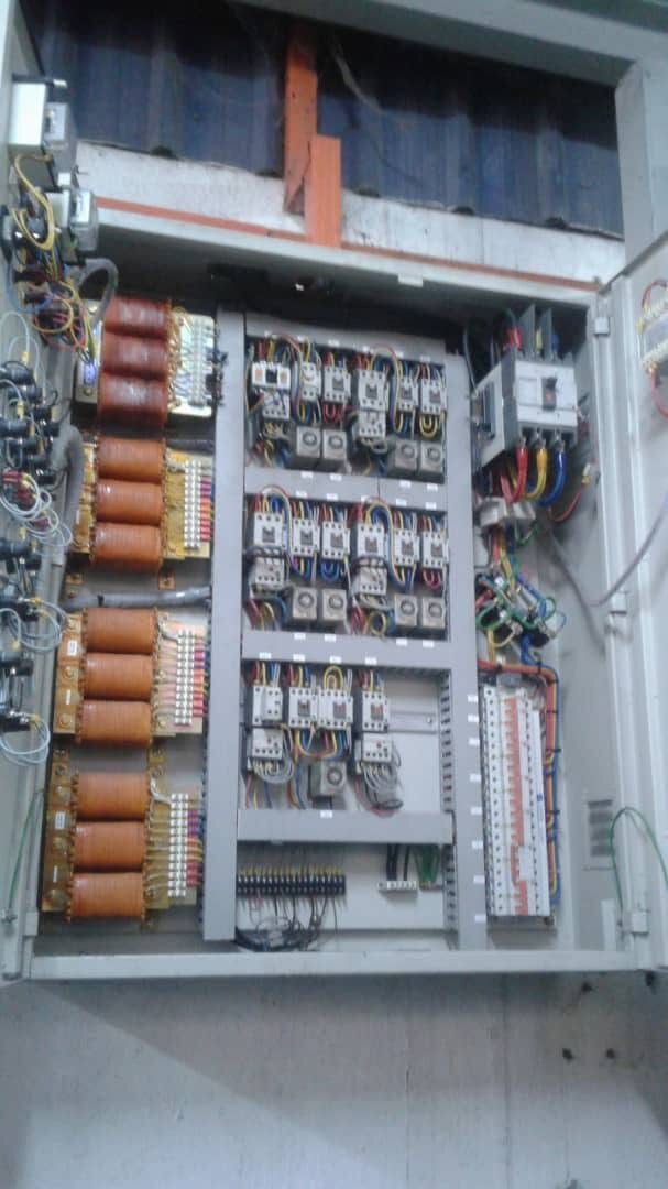 Grey control panel with its door open: four stacks of copper-coloured coils on the left, three rows of white contactors and relays with coloured wiring in the middle, a larger breaker and a row of small circuit breakers on the right