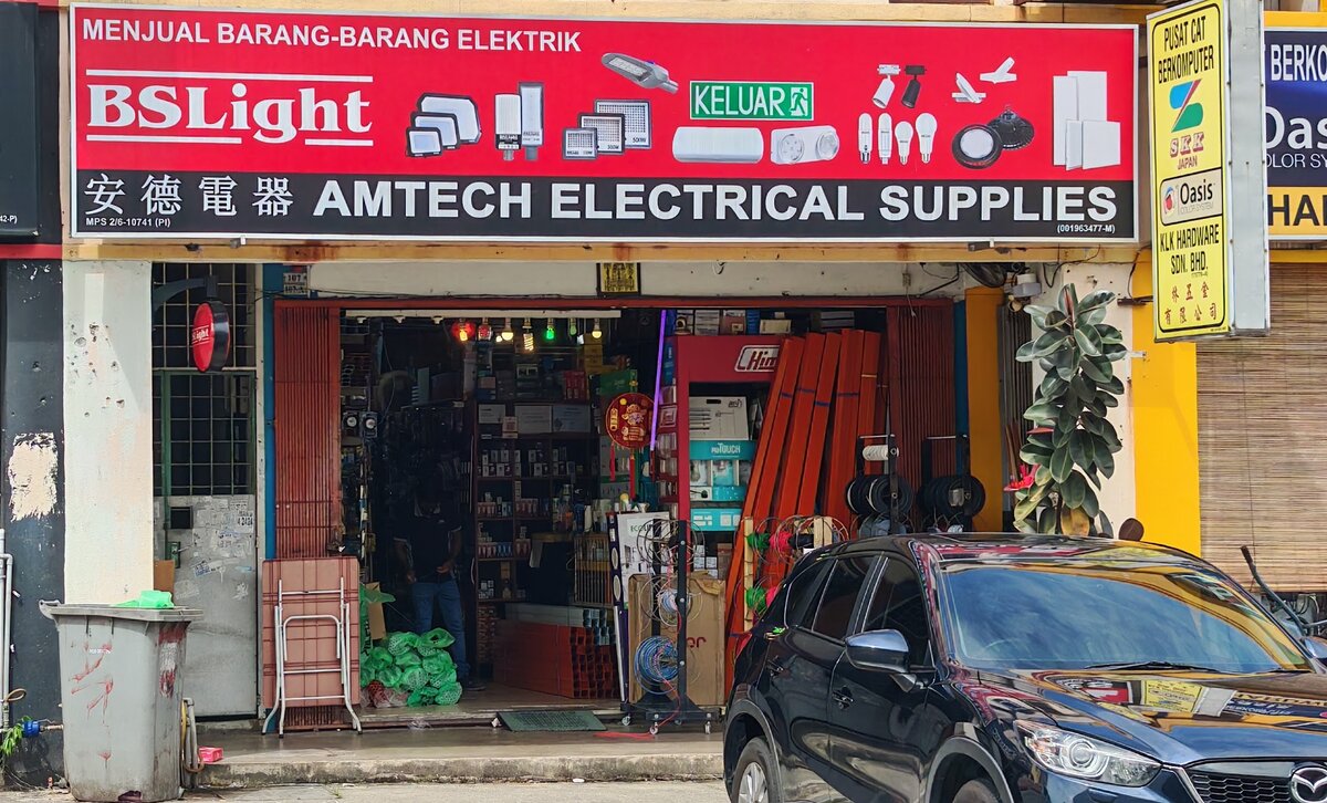 Red shop signboard reading Menjual Barang-Barang Elektrik, BSLight and Amtech Electrical Supplies with LED light pictures, above an open shopfront full of electrical goods, a red Himel frame, long orange lengths and cable reels