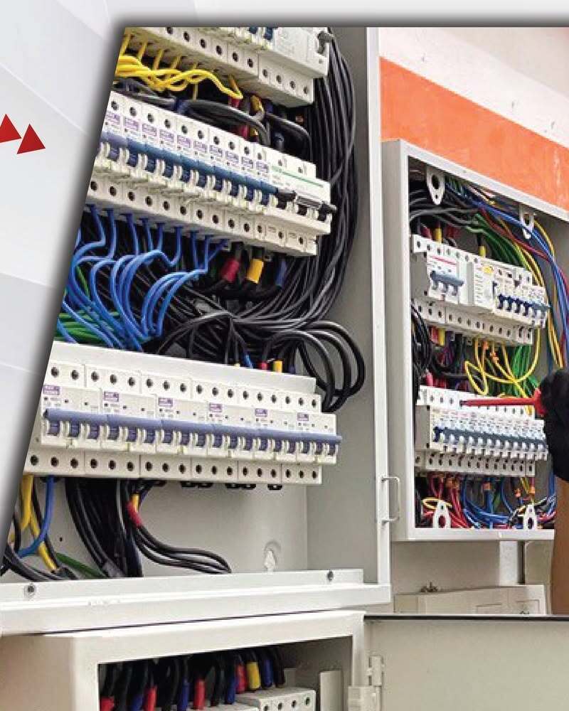 Two open grey distribution boards: the left one with rows of MCBs and bundles of blue, black and yellow cables, the right one smaller with MCBs and coloured wires, against a white and orange wall; a gloved hand at the right edge