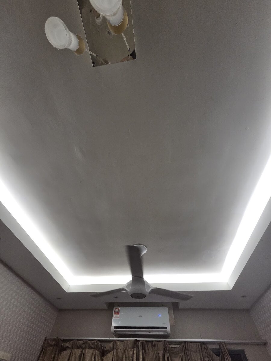 Ceiling with a recessed border lit by a cool white LED strip, a white ceiling fan in the middle and an aircond unit on the wall below