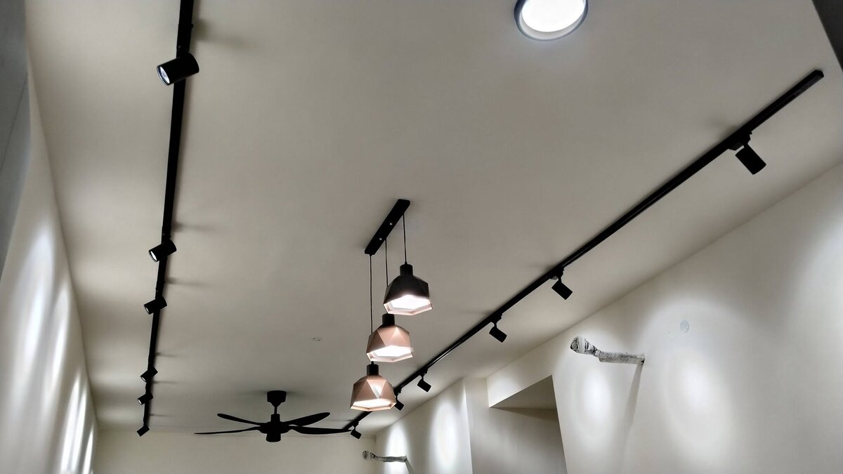 White ceiling with two long black track rails carrying spotlights, three geometric pendant lamps on one bar, a round flush ceiling light and a black five-blade ceiling fan