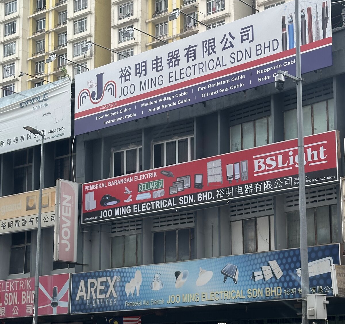 Row of shophouses under apartment towers with a large blue and white Joo Ming Electrical cable signboard, a red BSLight banner and a blue AREX Lightings banner