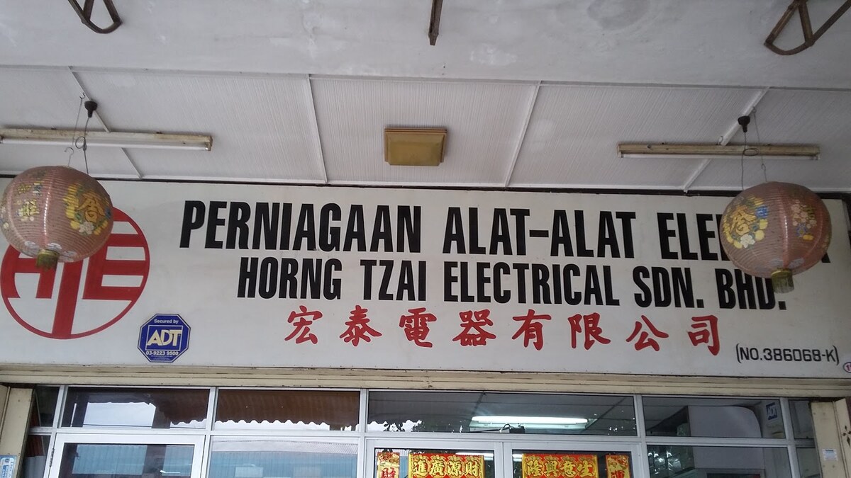 White shop signboard reading Perniagaan Alat-Alat Elek…, partly hidden by a lantern, Horng Tzai Electrical Sdn. Bhd., with red Chinese characters and a red HTE logo, between two paper lanterns