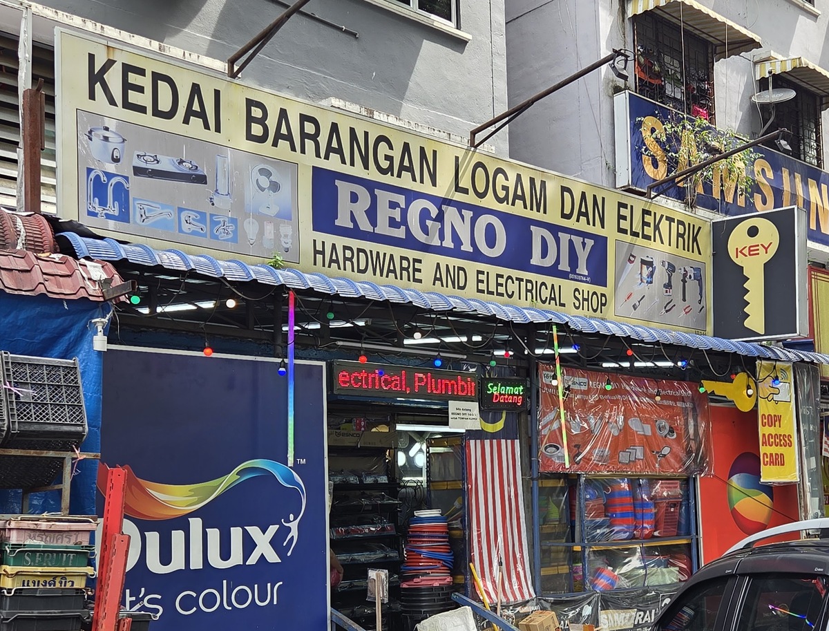 Yellow Regno DIY Hardware and Electrical Shop signboard with pictures of gas cookers, taps and tools, above an awning, a large Dulux panel and a key-shaped sign