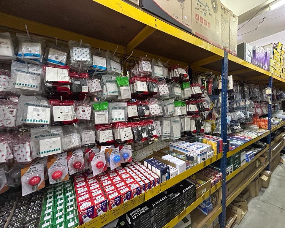 Shop aisle with walls of packed switches and sockets, coloured lamps, rows of boxed lamps and downlights, and boxed stand fans on the top shelf