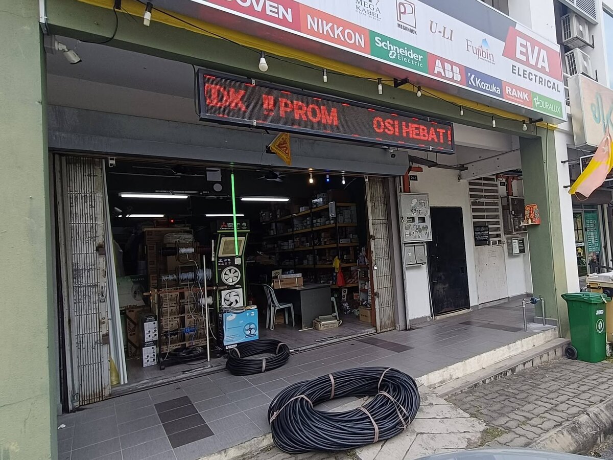 Open shopfront under a long white signboard with coloured brand panels and an EVA Electrical logo, a red LED ticker above the door and coils of black cable on the pavement