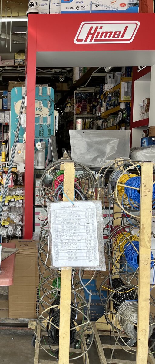 Shop entrance with a red Himel-branded display frame, shelves of electrical goods behind, and wooden stands holding reels of blue, yellow, red, black and grey cable with a printed price list in a plastic sleeve