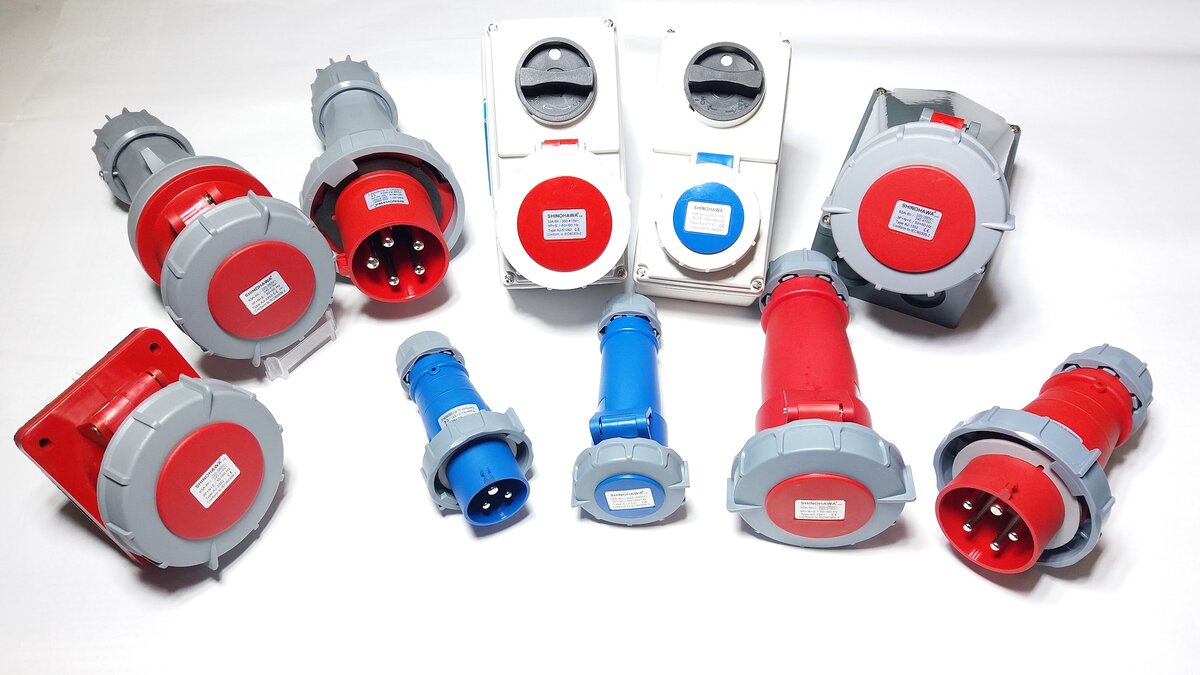 Red and blue industrial plugs, connectors, wall sockets and two switched socket boxes laid out on a white background