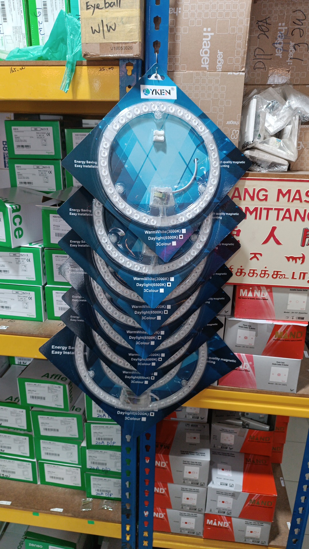 Shelves of green Schneider switch boxes and red Mand boxes, Hager cartons at the top and a column of packaged circular LED modules hanging in front