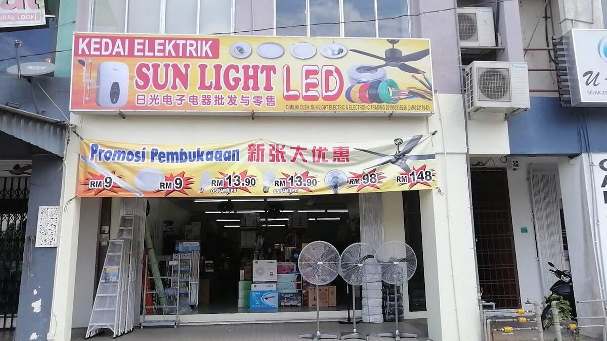 Shophouse with a yellow and red "Kedai Elektrik Sun Light LED" signboard, an opening-promotion banner with LED bulb and fan prices, and stand fans at the entrance