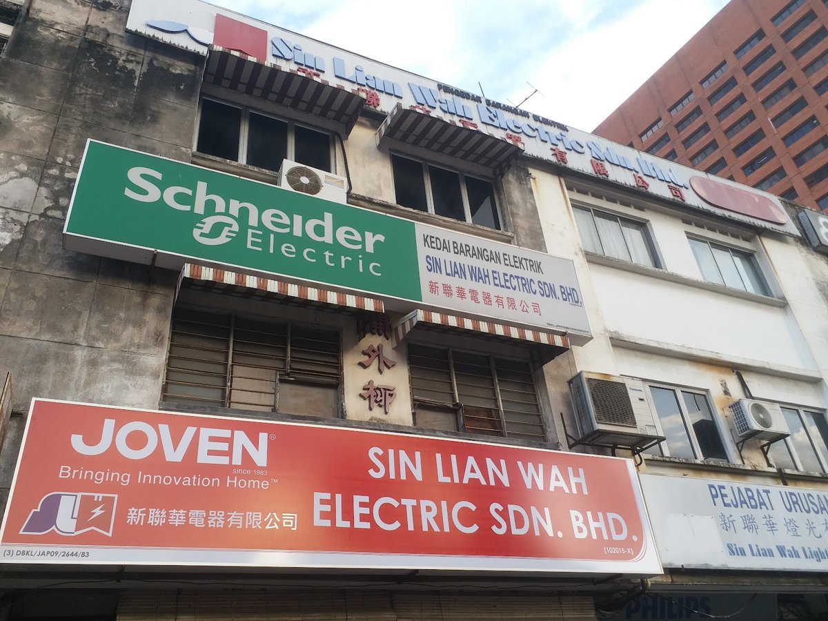 Shophouse front with a green Schneider Electric sign reading Kedai Barangan Elektrik Sin Lian Wah Electric Sdn. Bhd., a red Joven sign below, and a neighbouring unit signed Pejabat Urusan Sin Lian Wah Lighting