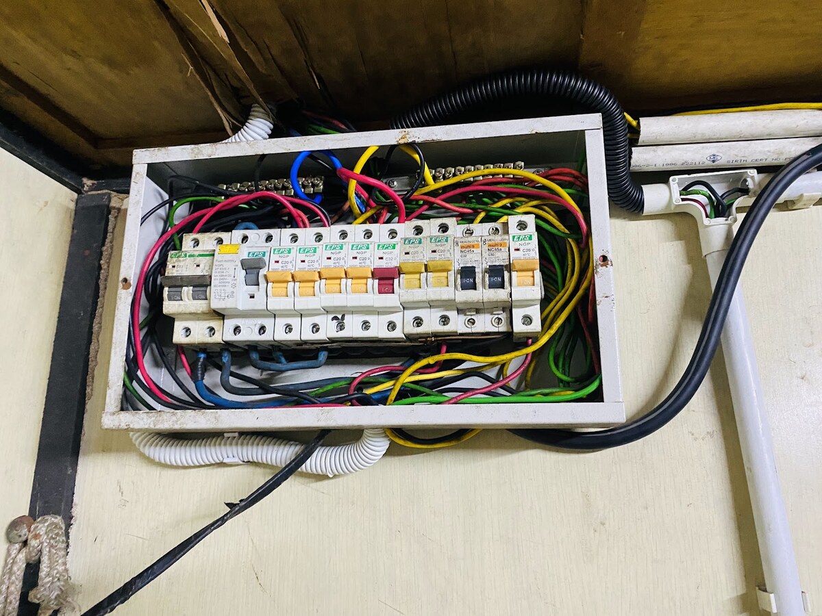 Open metal distribution box in a timber cabinet with a single row of breakers, most with EPS labels and two older black-toggle breakers, and coloured wires looping inside