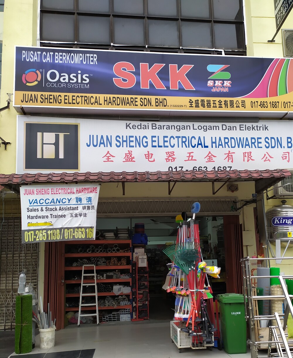 Shophouse with a dark blue SKK paint signboard and a white Juan Sheng Electrical Hardware signboard above an open front with shelves of pipes, a ladder, brooms and a green bin