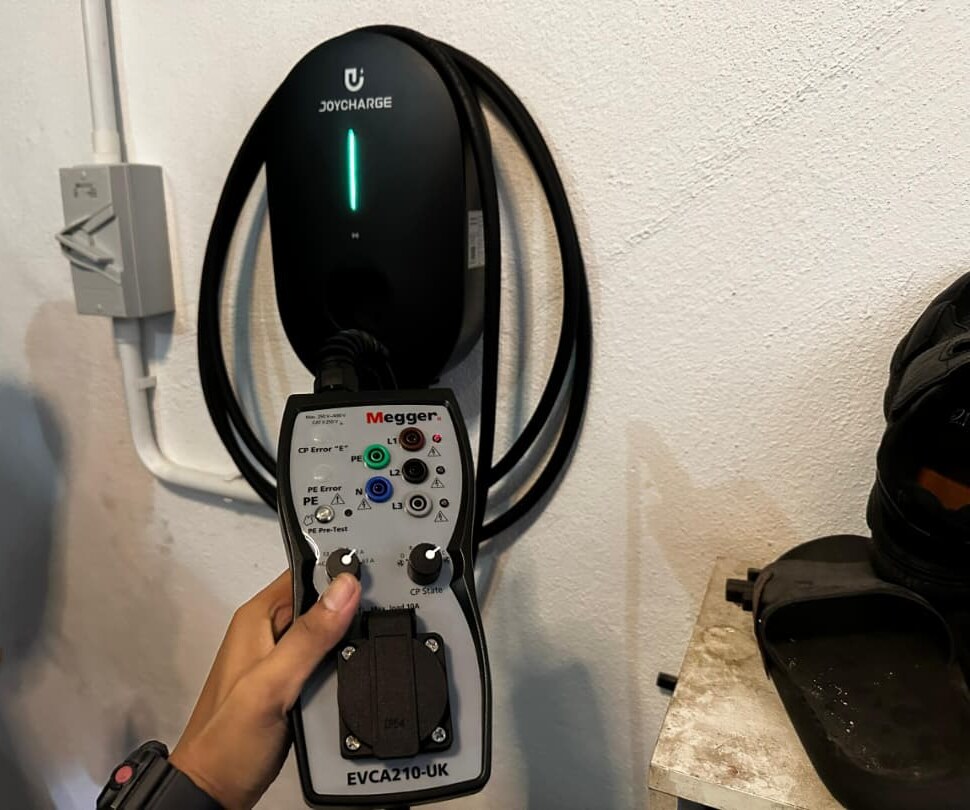 Black wall-mounted electric-car charger with a coiled cable and a green light, a hand holding a grey Megger test adapter plugged into it, and a wall isolator beside